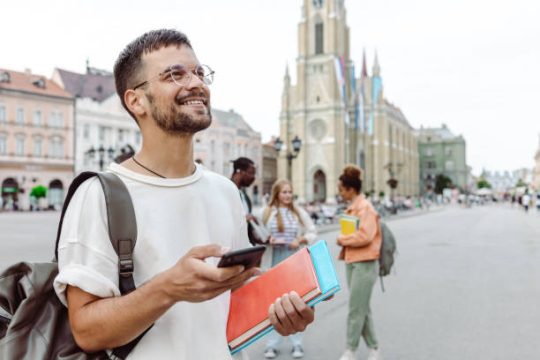 A young man carrying notebooks and using smart phone on the street while walking. A group of other students is behind