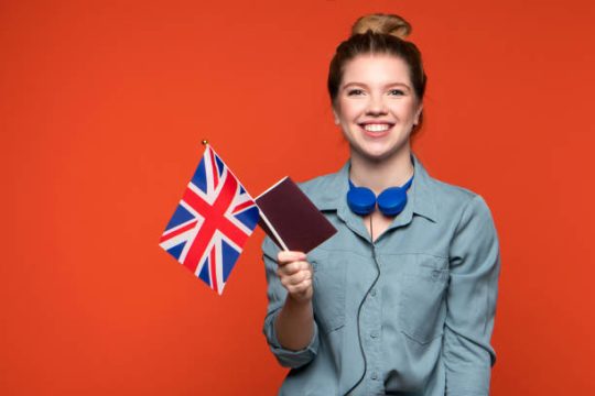 Young Smiling Woman in Casual Blue Shirt with UK Flag and Passport indoor orange background. Joyful Friendly looking exchange student girl looking at camera. Study abroad concept.