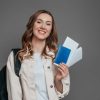 study abroad concept. student girl holding backpack, book, notebook, passport isolated on a dark grey background, copy space, immigration,