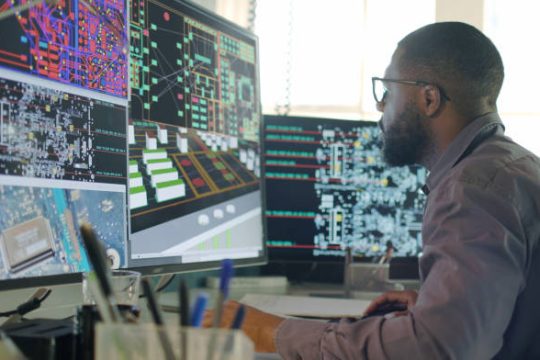 Stock image of an Afro-Caribbean male designing electronic circuit boards ( PCBs).
He’s sitting at a desk with a large computer screen displaying a variety of apps & PCB illustrations.