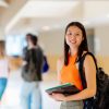 Portrait of an Asian girl with a backpack and school supplies in the hallways of the university campus with her classmates in the background.